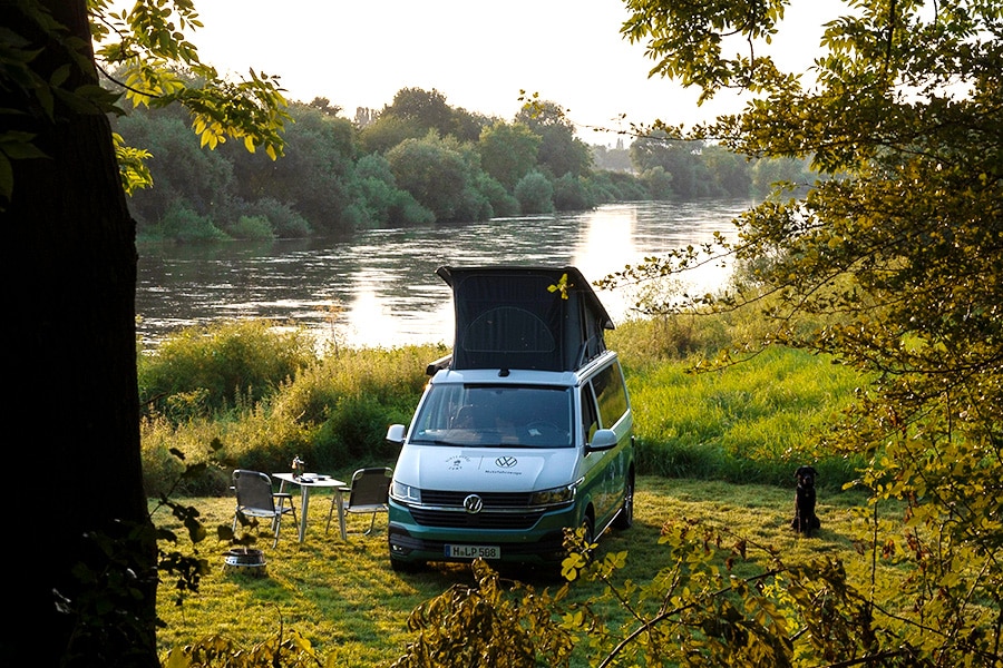 Campen mit einem VW Campingbus aud einer grünen Wiese am Fluss. Foto: Schau aufs Land