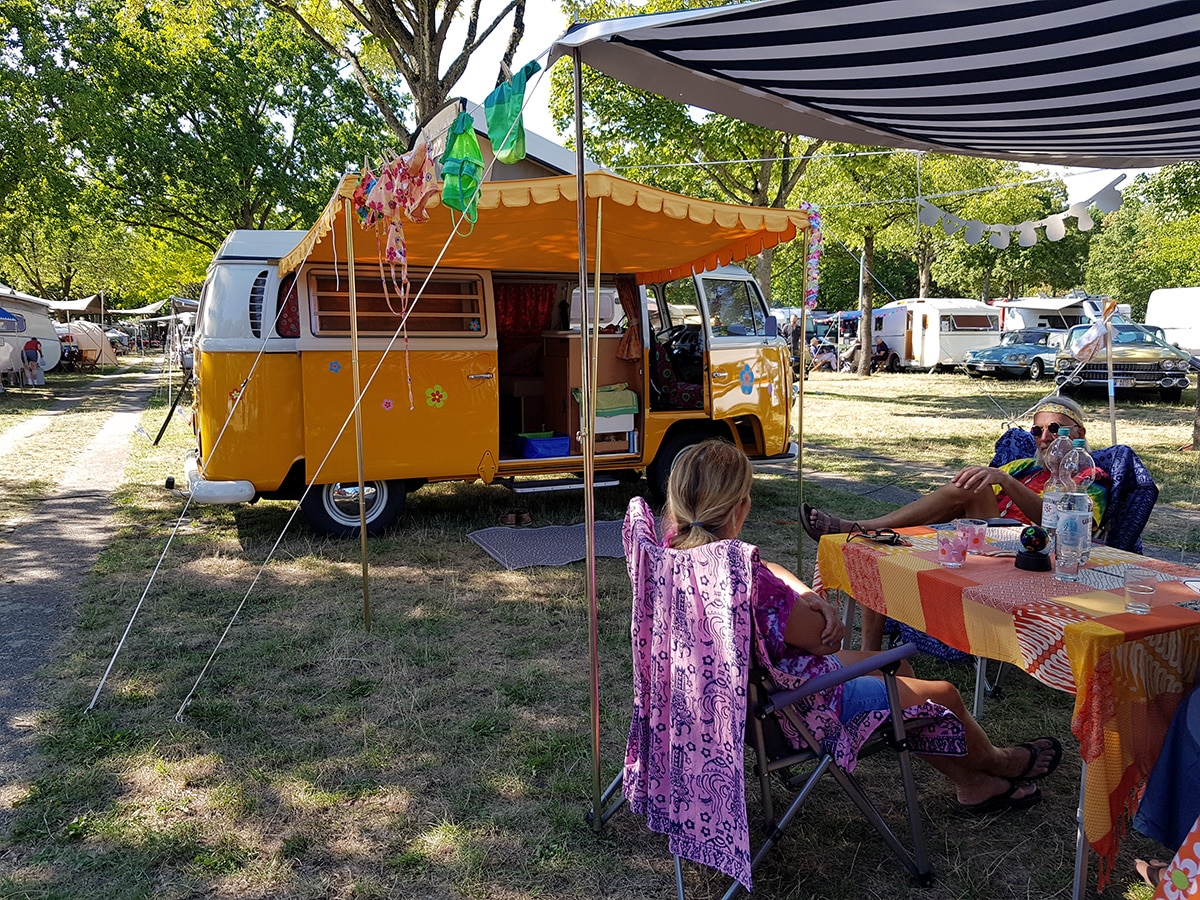 Campinggruppe im Hippie Stil vor gelb-weissem VW T2 Campingbus mit gelber Stoffmarkise