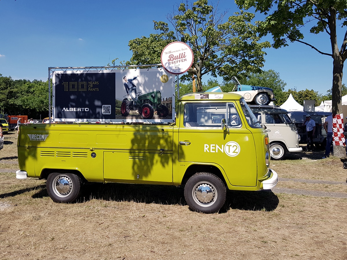 Ein moosgrüner VW T2 Pritschenwagen mit Werbeschild