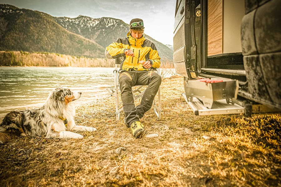Ein Camper sitzt mit seinem Hund vor dem Campingbus und grillt Fleisch auf einem mobilen Grill, der auf der Trittstufe des Fahrzeugs steht