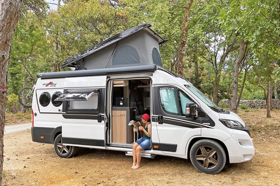 Malibu Van Diversity 640 LE K zwischen Pinienbäumen, mit Camperin und Hund an der offenen Schiebetür