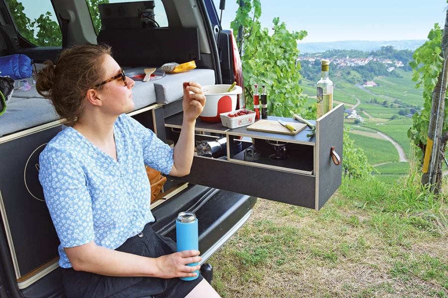Heckbereich mit ausgezogenem Küchenblock und daneben sitzender Camperin