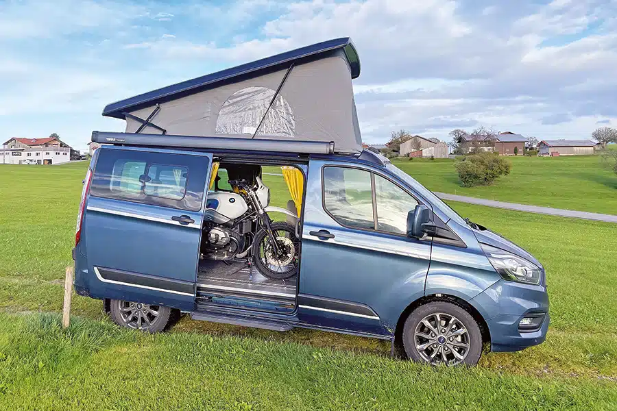 Mittelblauer Campingbus Southvan Allrounder mit Aufstelldach und mit Motorrad im Innenraum auf einer gemähten Wiese