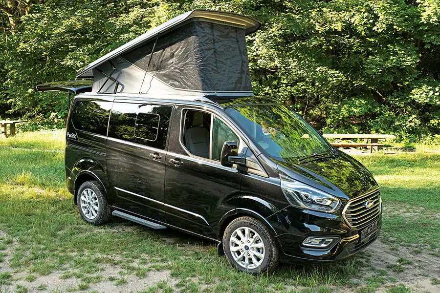 Schwarzer Campingbus Vanestro Pacific mit Aufstelldach auf Wiese am Waldrand
