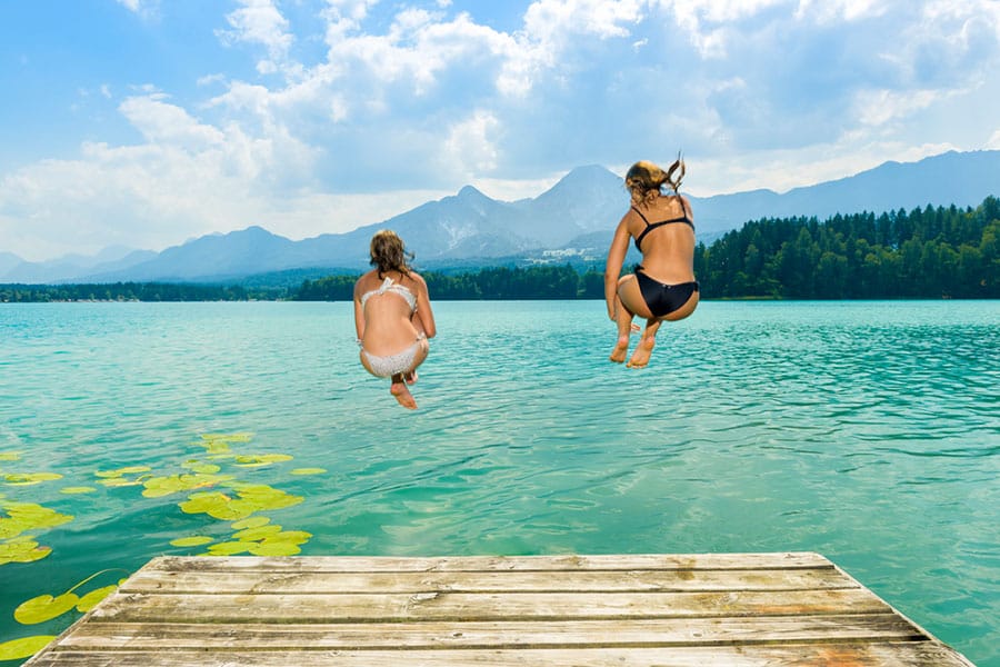 Zwei Mädchen springen von Holzsteg ins türkisblaue Wasser eines Sees am Fuß der Alpen.