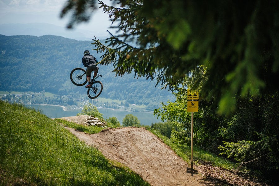 Bucketride Festival: Mountainbiker während eines Sprungs auf hügeliger Trail-Piste mit See- und Bergkulisse im Hintergrund