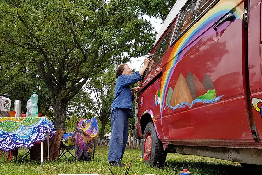 Frau bemalt mit Pinsel einen roten VW T3 Oldtimer im Hippie-Stil