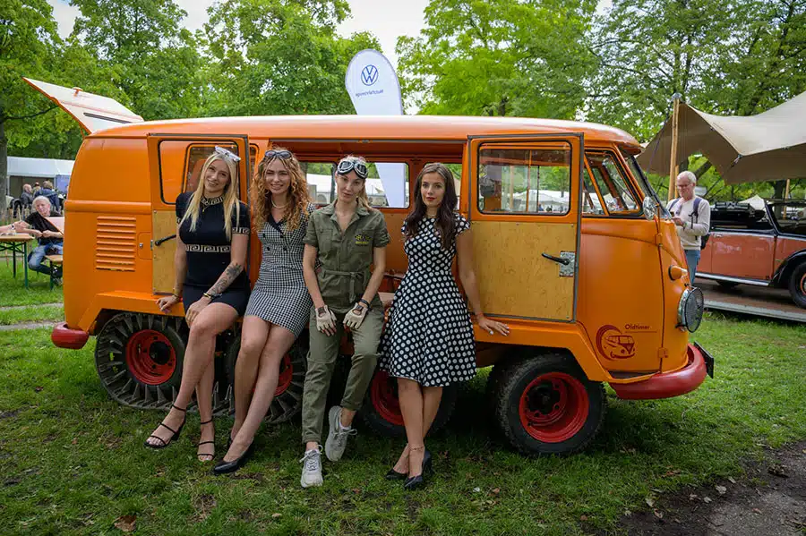 Vier junge Frauen sitzen im seitlichen Türeingang eines orangefarbenen VW Bus Oldtimers
