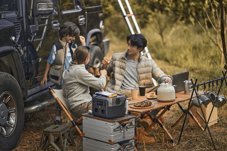 Camping-Familie sitzt draußen am Tisch, daneben mobile Powerstation mit angeschlossenen Geräten zum Aufladen