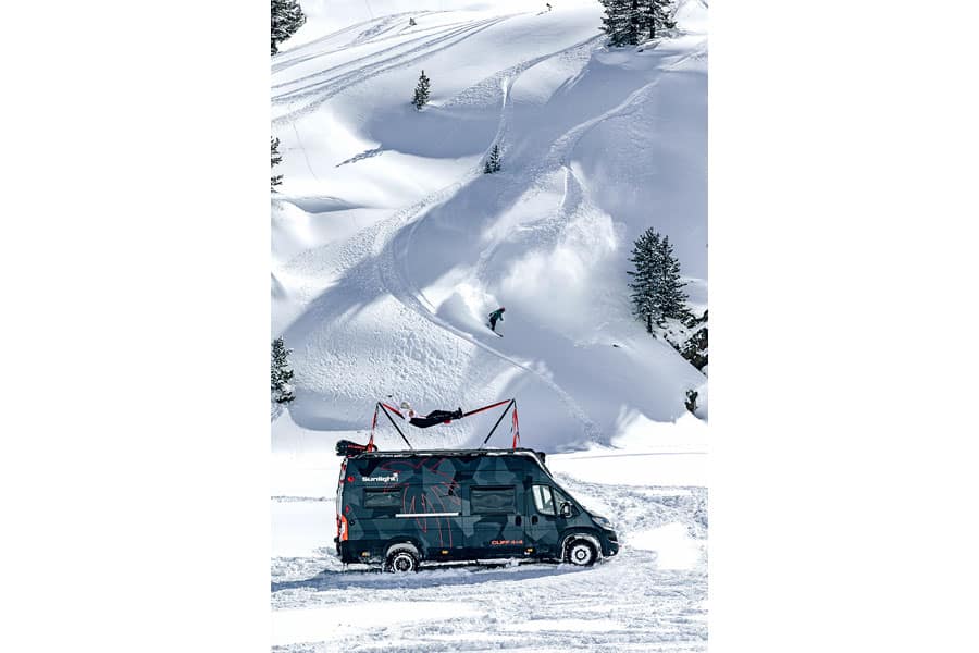 Sunlight Cliff 4x4 Adventure Van im Schnee vor einem Berghang mit Snowboardpiste, auf der ein Snowboarder herunterfährt