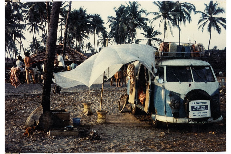 Blau-weißer T1 Bulli mit Dachgepack und weißem Sonnensegel auf dem Strand, im Hintergrund Menschen, ein kleines Strandhaus und Palmen