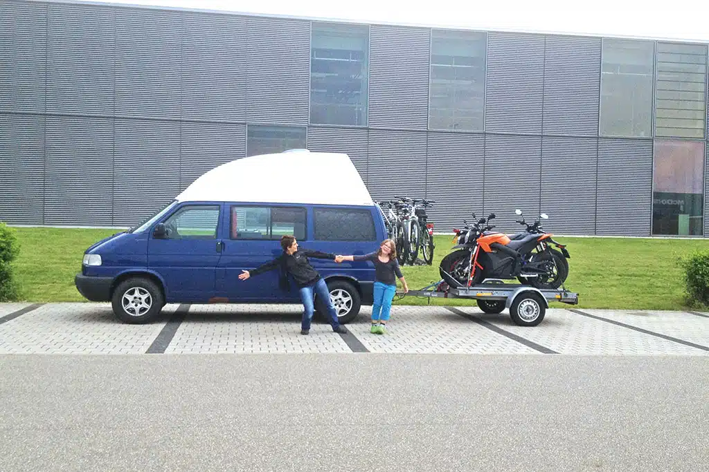 Blauer VW-Campingbus mit Fährrädern am Heckträger und Motorrädern auf kleinem Anhänger, vor dem Camper stehen eine Frau und ein Mädchen