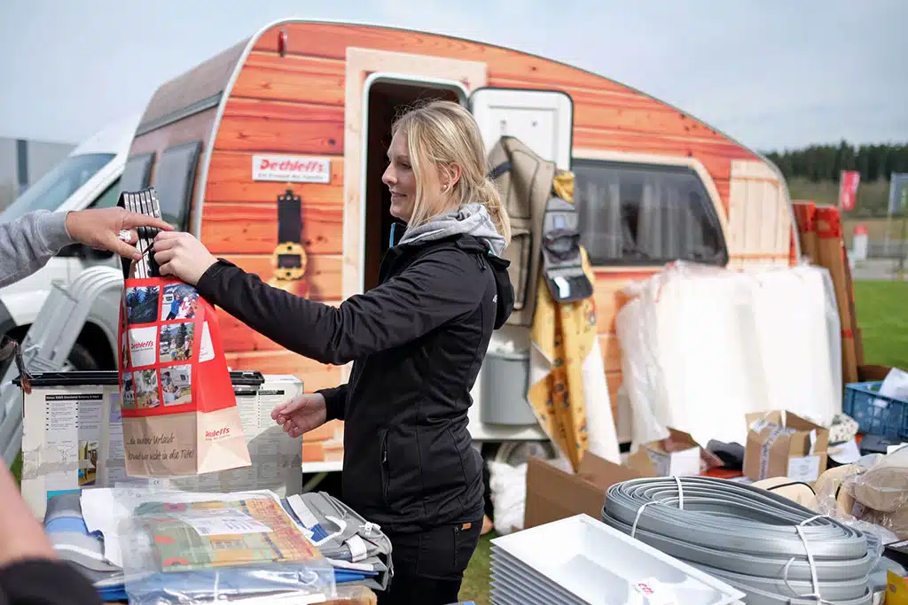 Frau bietet Gebrauchtes auf Camping-Flohmarkt an, im Hintergrund ein alter Wohnwagen