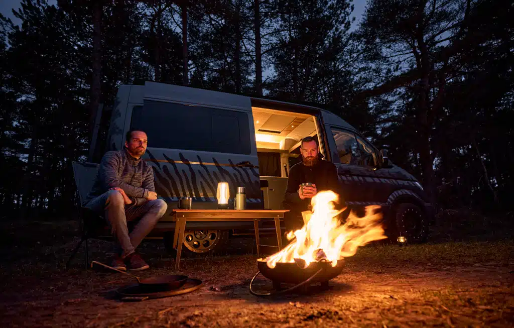 Mann sitzt bei Nacht vor einem Campervan, dessen seitliche Schiebetür geöffnet ist, vor dem Mann brennt ein Feuer in einer Feuerschale