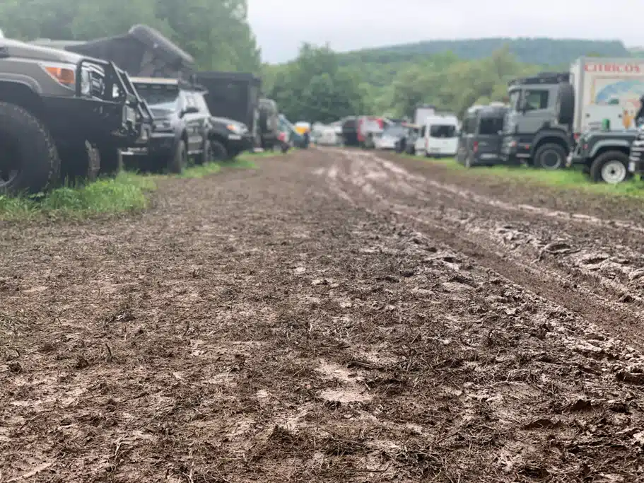 Expeditionsfahrzeuge und 4x4-Campingbusse auf einer Wiese mit viel Baumbestand, zwischen den Fahrzeugreihen befindet sich ein verschlammter Weg