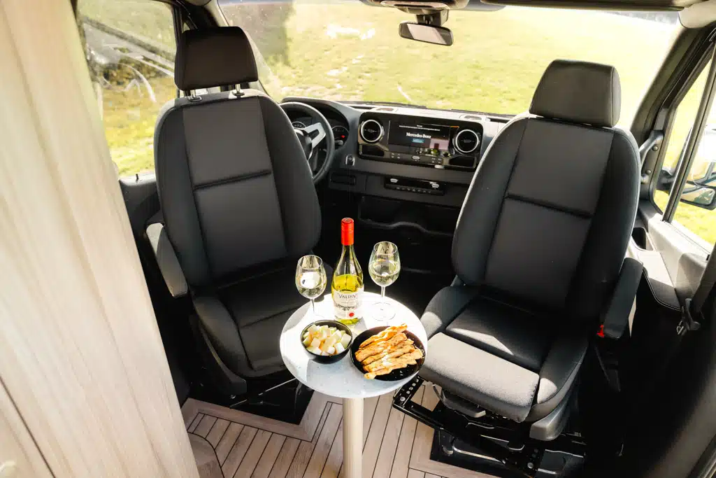 Um rund 90 Grad gedrehte Vordersitze im Kastenwagen, davor ein kleiner, runder Tisch mit Weinflasche, zwei Gläsern und zwei Schalen mit Snacks