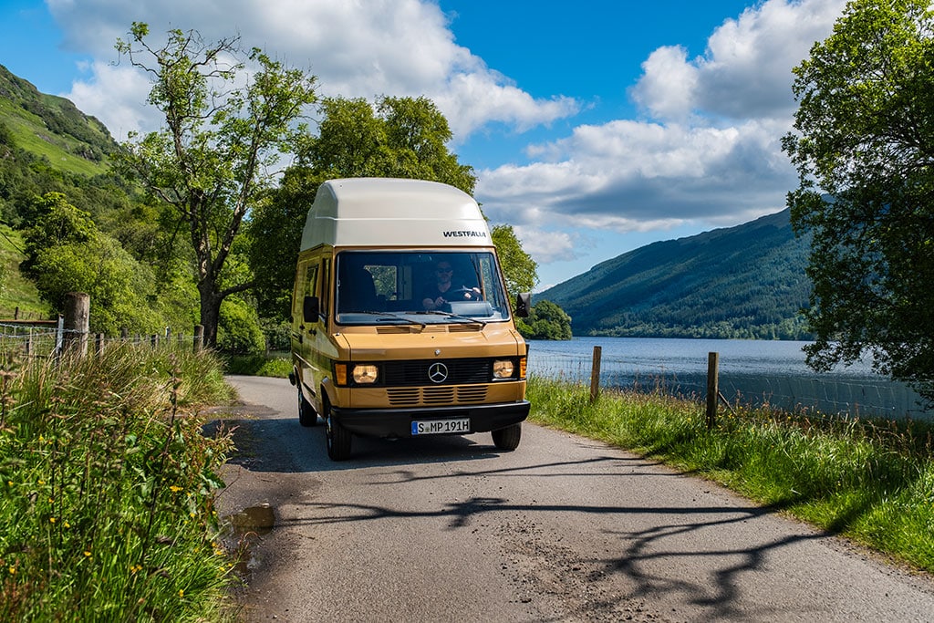 Alter Mercedes Marco Polo- Campervan auf einer Landstraße an einem See in einer Hügellandschaft