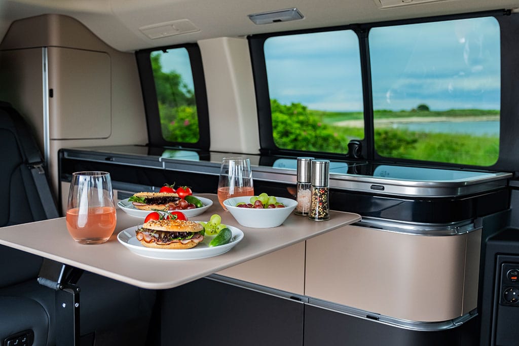 Küchenmodul im Campervan mit ausgeklapptem Tisch und zwei Tellern mit Burgern sowie mehreren gefüllten Gläsern
