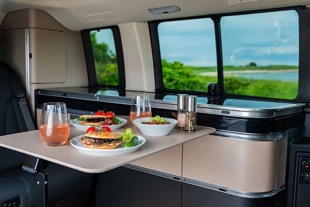 Küchenmodul im Campervan mit ausgeklapptem Tisch und zwei Tellern mit Burgern sowie mehreren gefüllten Gläsern