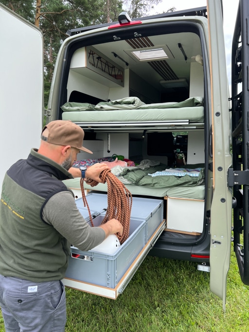 Mann holt einen Gegenstand aus einer Eurobox, die auf einem Schwerlast-Heckauszug am Campervan steht