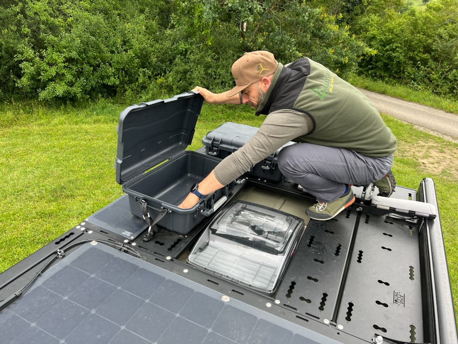 Mann hockt auf der Dachterrasse eines Campervans und öffnet eine dort befestigte, schwarze Box