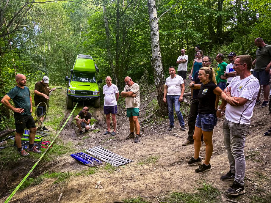 Teilnehmer eines Workshops stehen um eine Offroadfahrzeug herum, das an einem Bergeseil hängt, davor liegen (Sand-)Bleche auf dem Boden