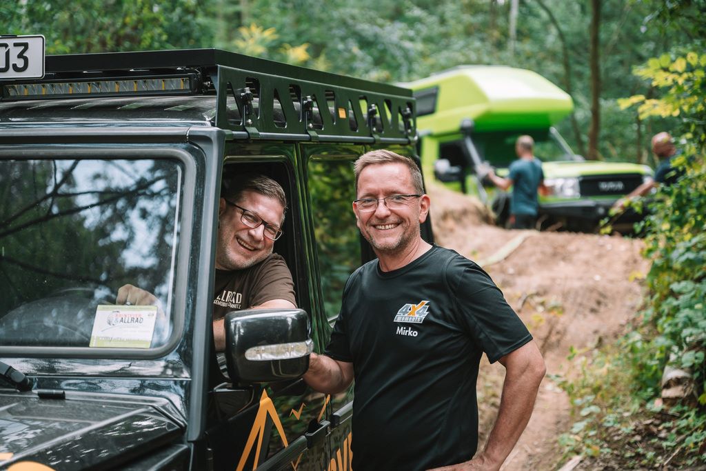 Zwei Männer in beziehungsweise neben einem Landrover lachen in die Kamera.