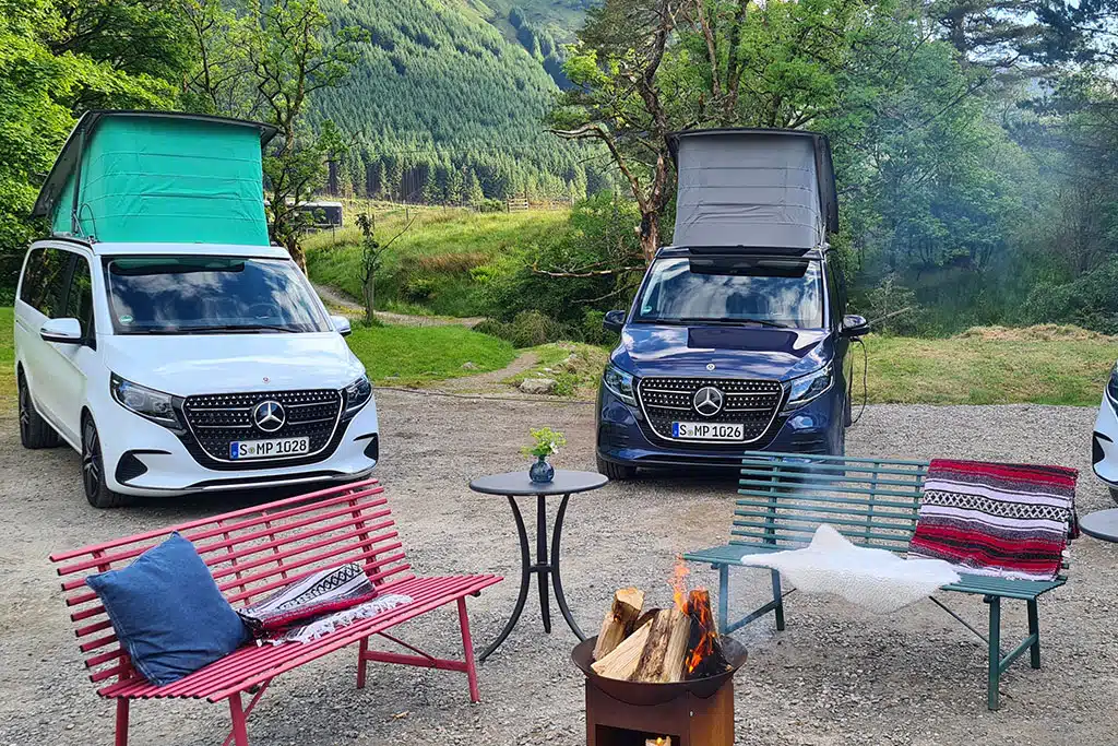Ein dunkelblauer und ein weißer Campervan mit Aufstelldach in grüner Landschaft, davor zwei Bänke, ein kleiner Tisch und eine Feuerstelle
