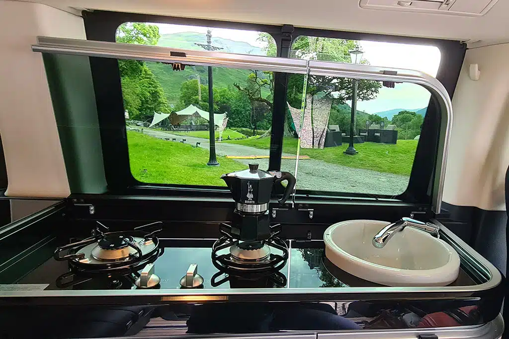 Küchenmodul im Campervan mit Ausblick durch das davor liegende Fenster auf eine grüne Hügellandschaft