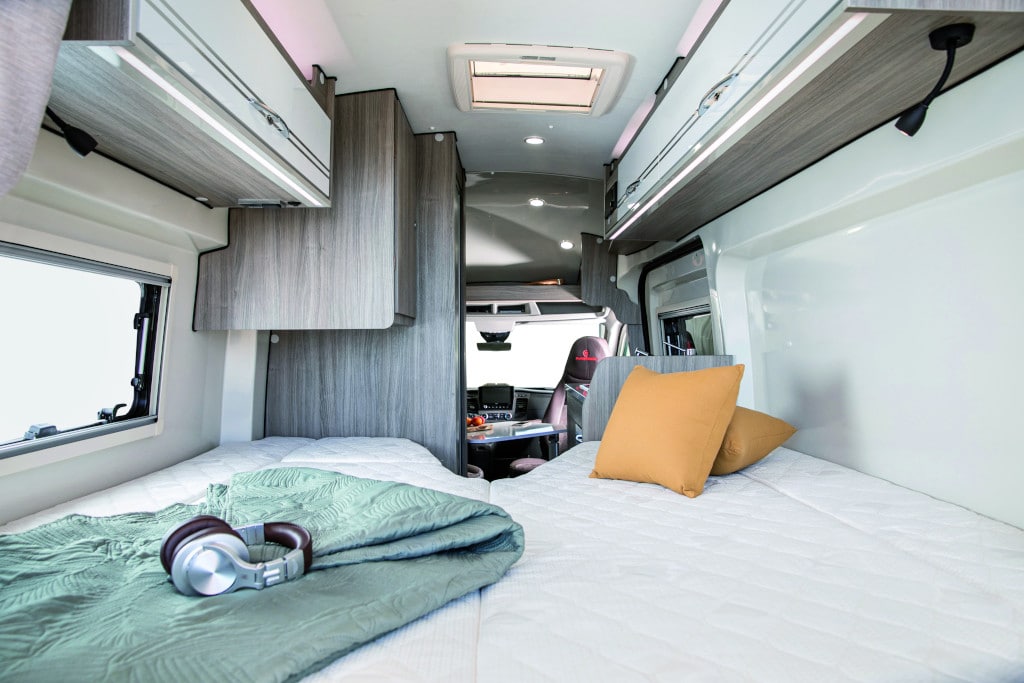 Blick durch die offene Hecktüre auf zwei zu einer Fläche zusammengefügten Längsbetten vor Dusch-Schrank und Fahrerkabine