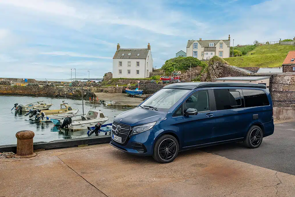 Dunkelblauer Marco Polo Campervan am Hafen, im Hintergrund eine Bucht mit kleinen Booten, dahinter das Ufer mit einigen Häusern