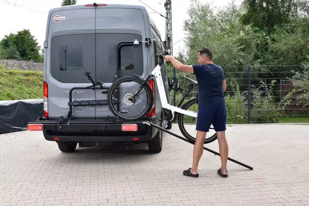 Ein Mann schiebt ein Fahrrad über eine Auffahrschiene auf einen Fahrradträger, der an einem Kastenwagenheck befestigt ist.