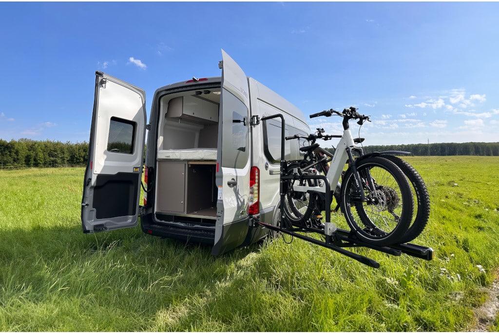 Ein Kastenwagen steht mit geöffneten Hecktüren auf einer Wiese, ein mit zwei Rädern beladener Fahrradträger ist komplett zur Seite geschwenkt, sodass der Zugang zum Heck frei ist.