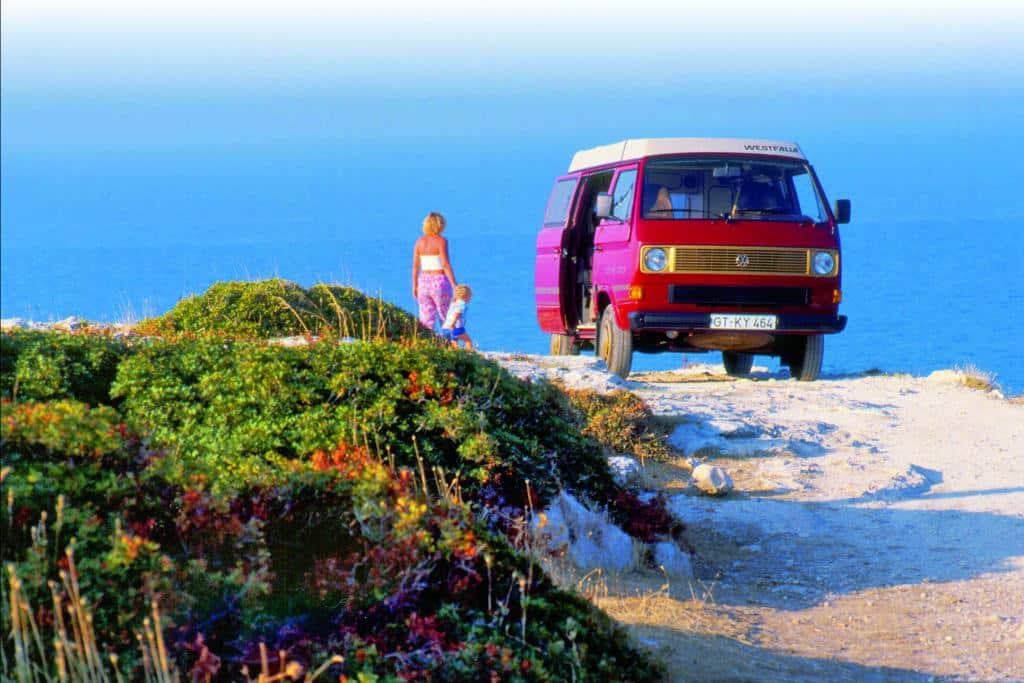 Altes Foto mit einem roten VW Westfalia Joker Campingbus am Meer, davor steht eine Frau mit einem Kleinkind an der Hand