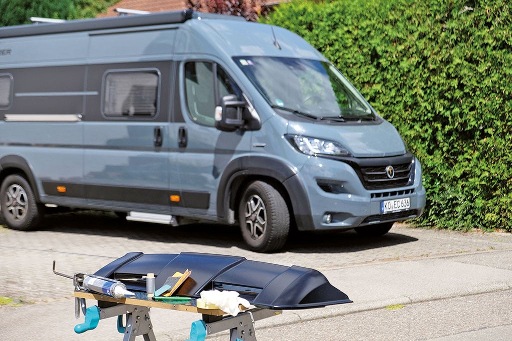 DIY Dachspoiler nachrüsten: Grauer Kastenwagen neben einer grünen Hecke, davor ein Dachspoiler und Klebe-Materialien zur Nachrüstung