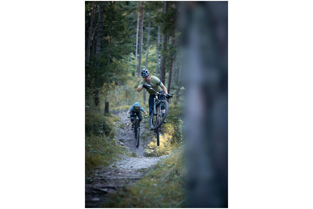 Ein Mountainbikefahrer beim Sprung mit seinem Bike auf einem gewundenen Trailweg im Wald, dahinter fährt ein weiterer Biker
