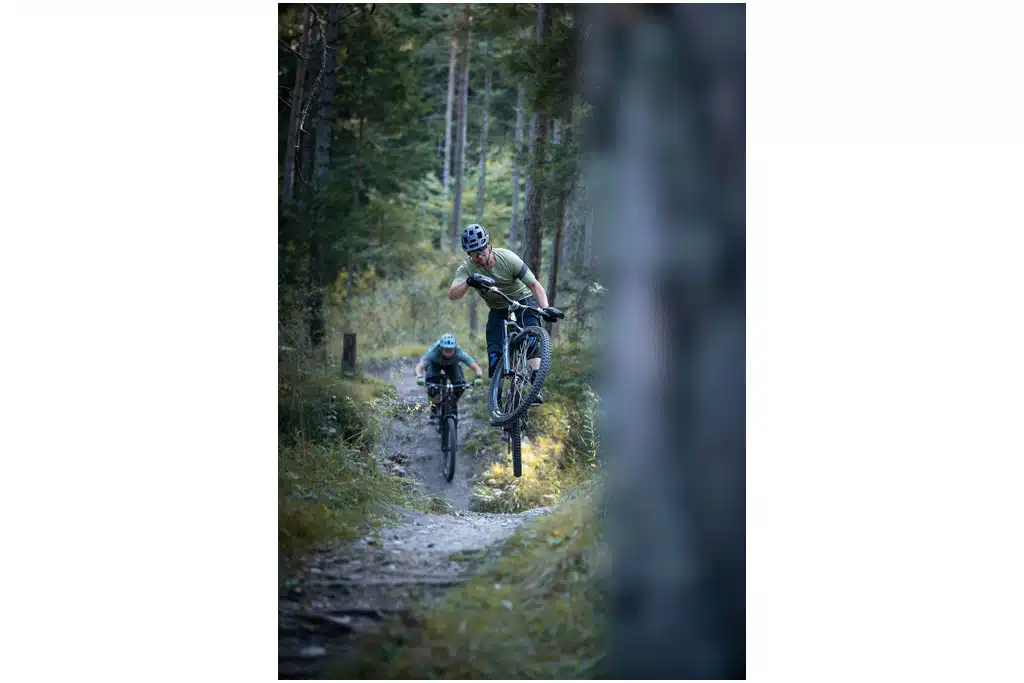 Ein Mountainbikefahrer beim Sprung mit seinem Bike auf einem gewundenen Trailweg im Wald, dahinter fährt ein weiterer Biker