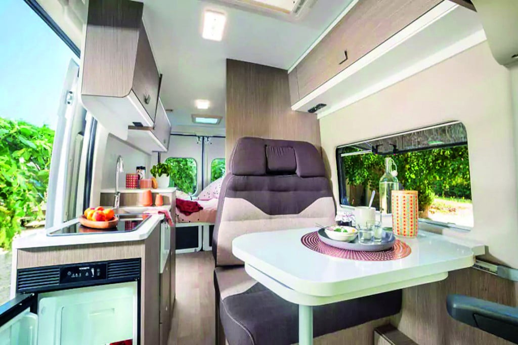 Blick nach hinten auf Sitzgruppe und Küchenblock geegenüber mit hellen Oberflächen, Polster dunkelbraun, helles Holzdekor der Möbel