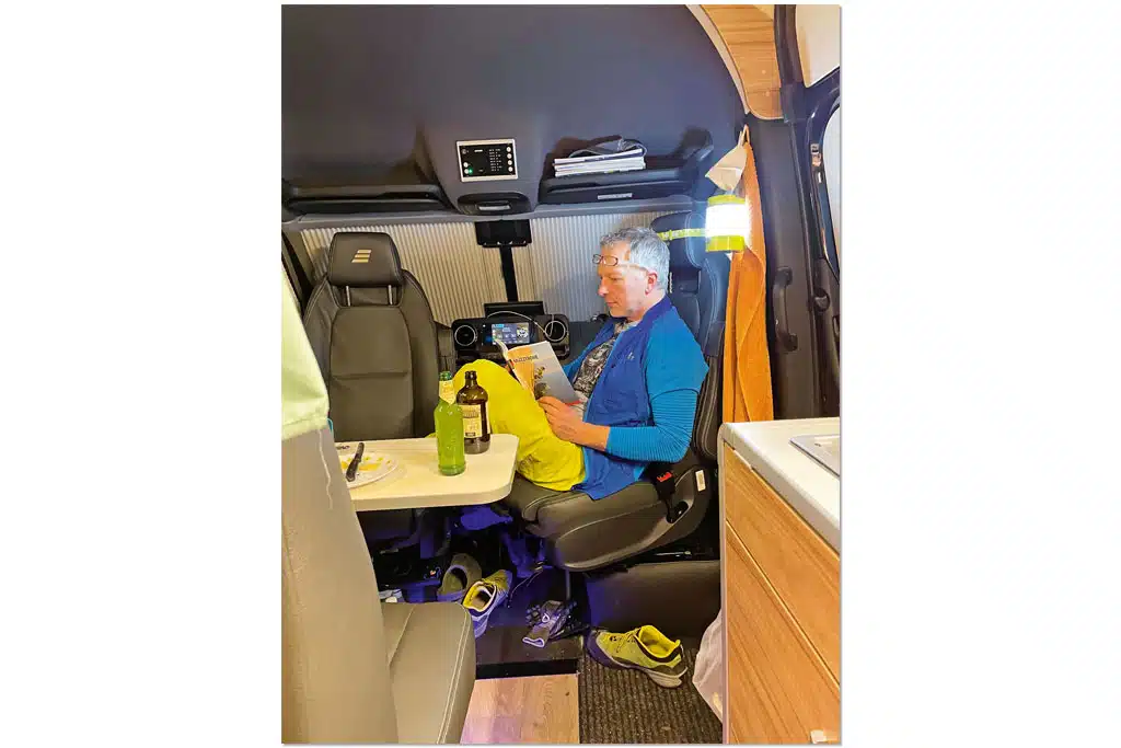 Mann sitzt ein Buch lesen auf dem umgedrehten Beifahrersitz eines Campervans
