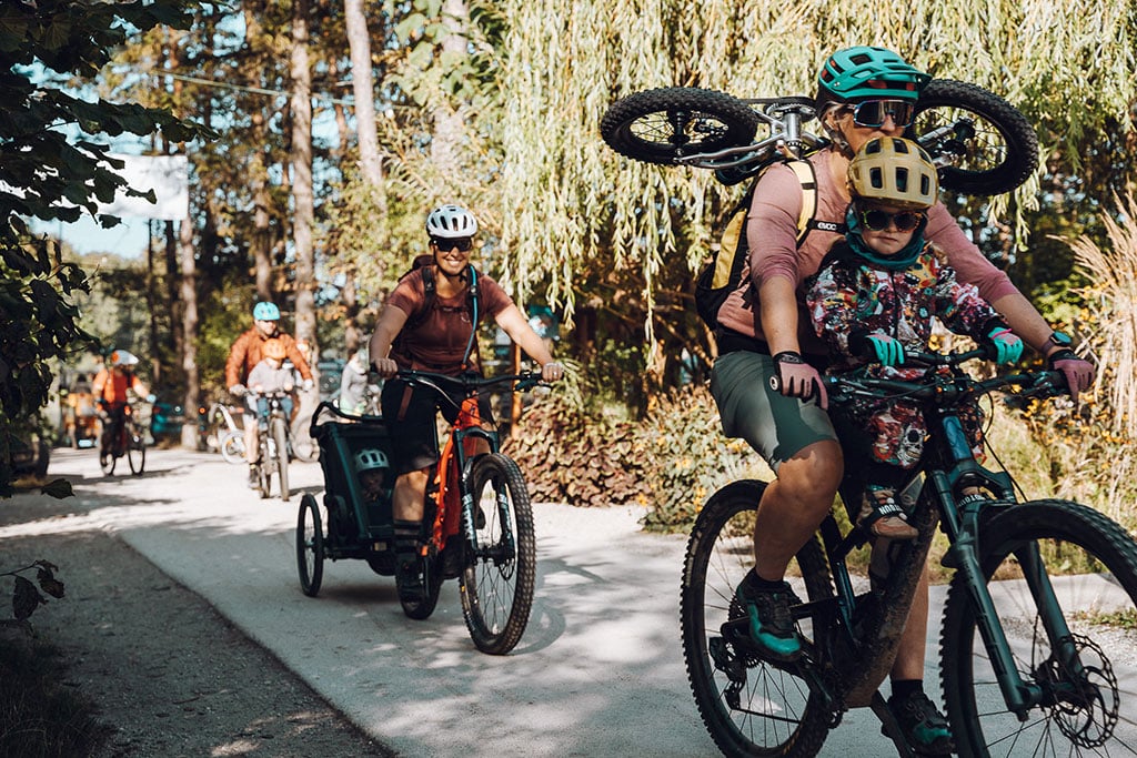 Ein Mann auf einem Mountainbike fährt auf einem schmalen Weg, auf dem Rad vor ihm sitzt ein Kleinkind, der Fahrer hat ein Kinder-Fahrrad auf seinem Rücken befestigt. Dahinter fahren noch weitere Mountainbiker und Mountainbikerinnen