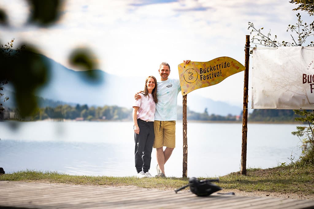 Eine Frau und ein Mann stehen neben einem auf einem Pfosten befestigten Wimpel mit Aufschrift Bucketride Festival am Seeufer