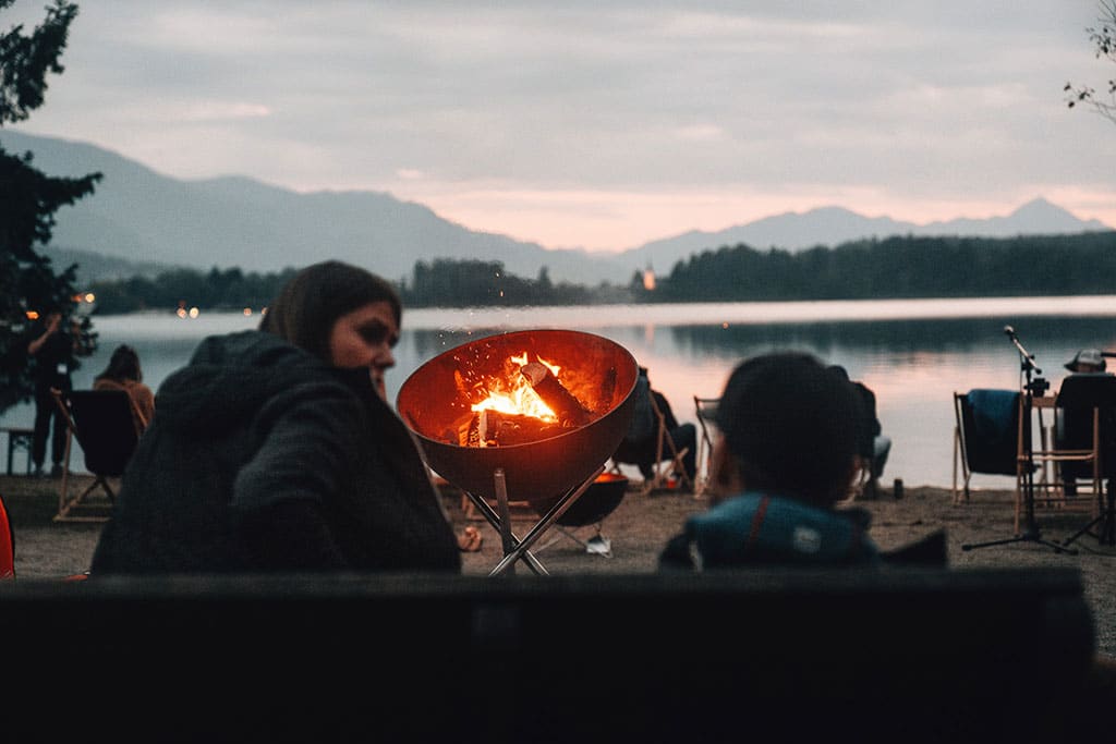 zwei Menschen sitzen am See, vor ihnen brennt ein Lagerfeuer