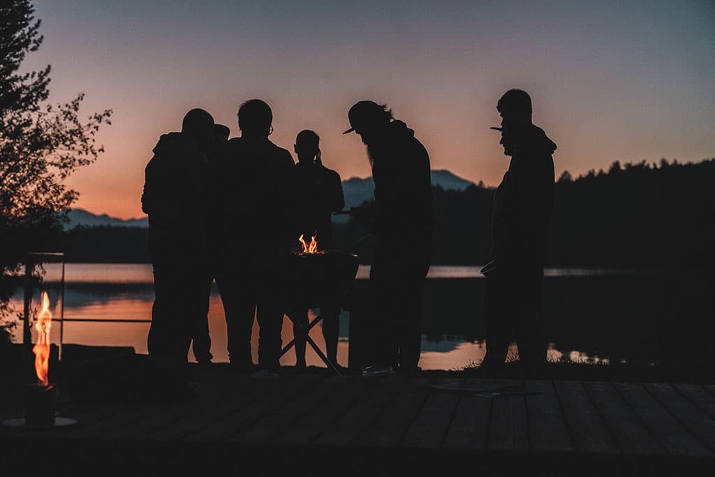 Mehrere Menschen stehen um ein kleines Feuer herum am Ufer eines Sees