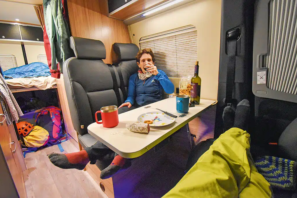Frau sitzt auf Rücksitzbank im Campervan, auf dem Tisch davor stehen eine Flasche Rotwein und Snacks