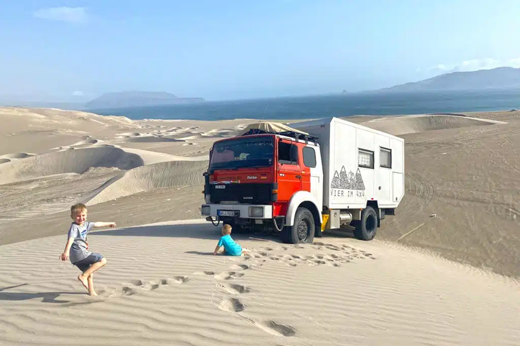 Zwei Kinder spielen in den Sanddünen einer Wüste, hinter ihnen steht ein rot-weißes Expeditionsmobil