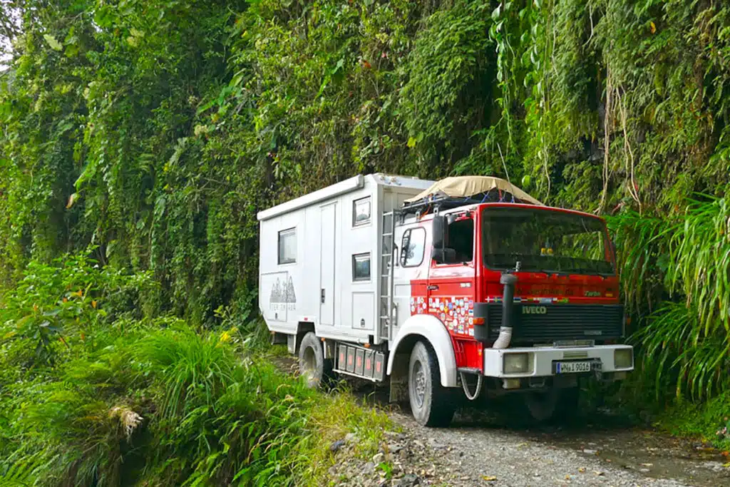 Expeditionsmobil mit weißer Wohnkabine und rotem Chassis auf kleiner, unbefestigter Straße an einem Berghang