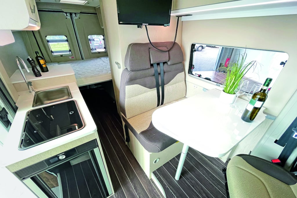 Zweier-Sitzbank mit Polster braun und beige; helle Oberflächen, helles Möbeldekor, dunkelbrauner Fußboden, Querbett im Heck