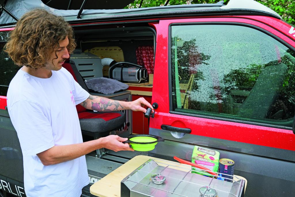 Mann zapft Wasser aus einem an der Fahrertür eines rot-grauen Campingbusses per Magnethalter befestigten Wasserschlauchs in einen hellgrünen Hundenapf
