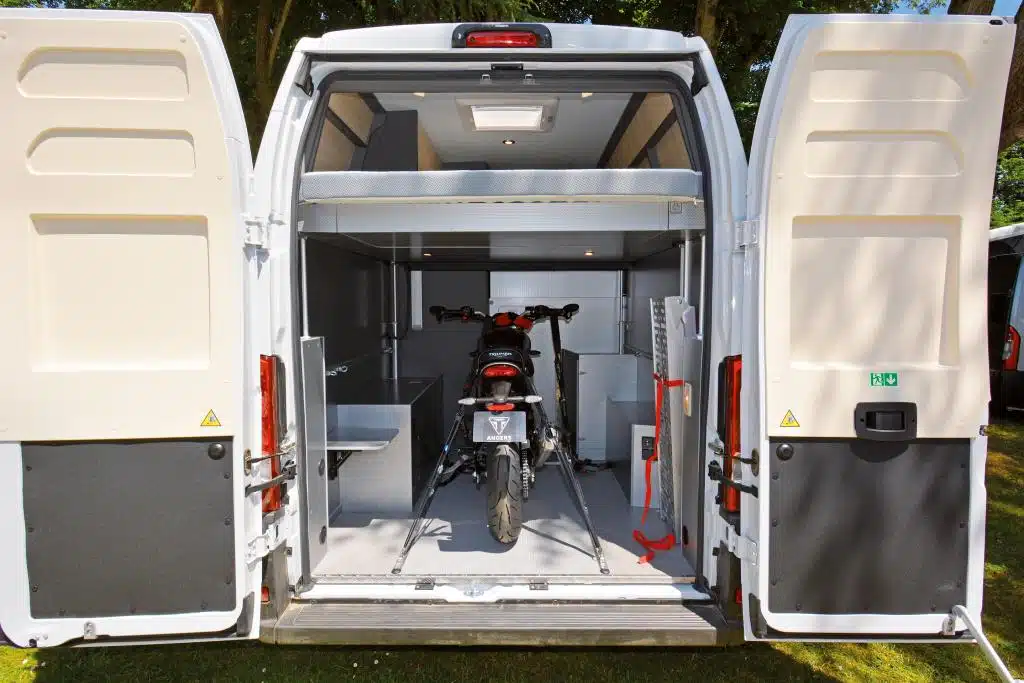 Campervan-Heckansicht bei offenen Hecktüren, unter dem unter die Decke hochgezogenen Hubbett steht mit Gurten fixiert ein Motorrad