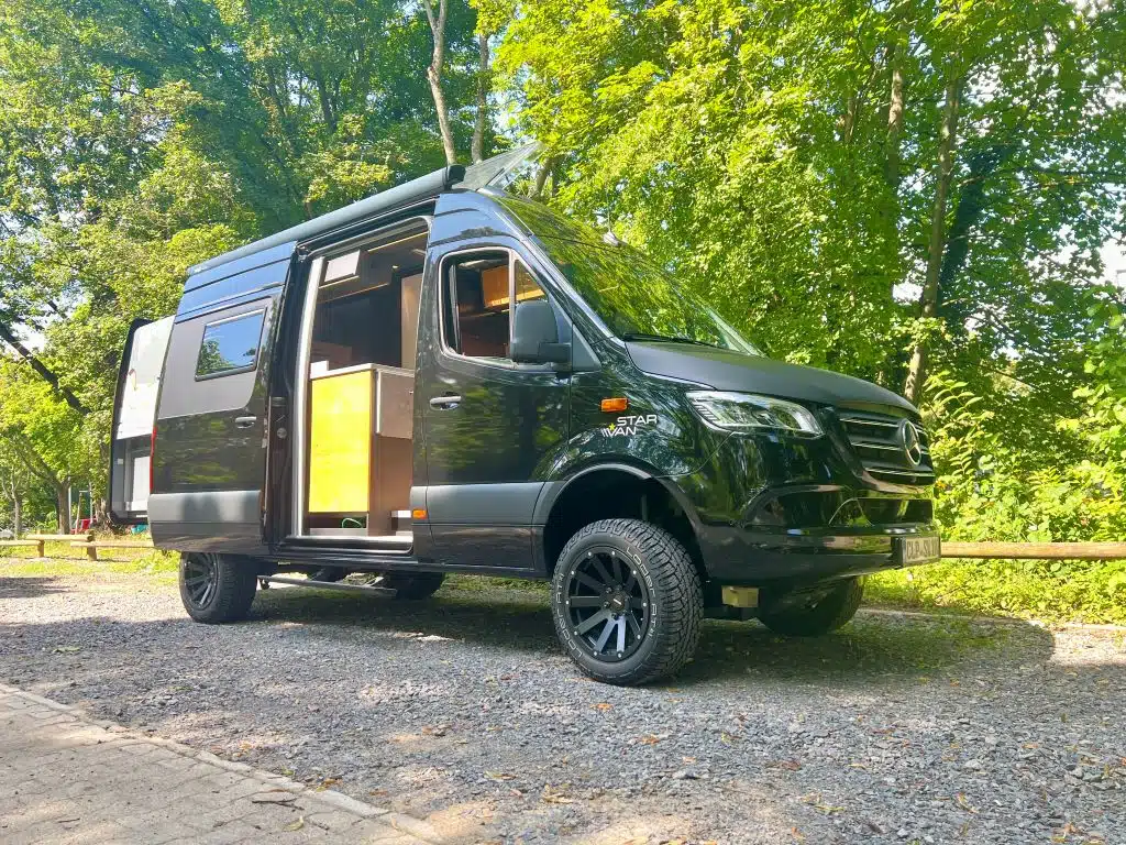 Schwarzer Kastenwagen-Campervan seitlich fotografiert vor einer Reihe Bäume